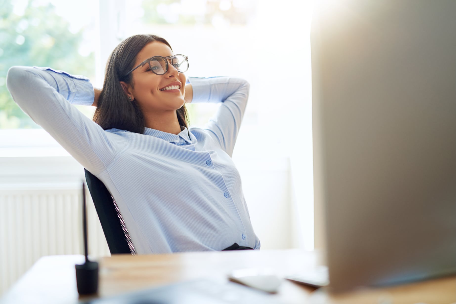 Smiling woman wearing  sits back relaxed on her desk, with her hands behind her back